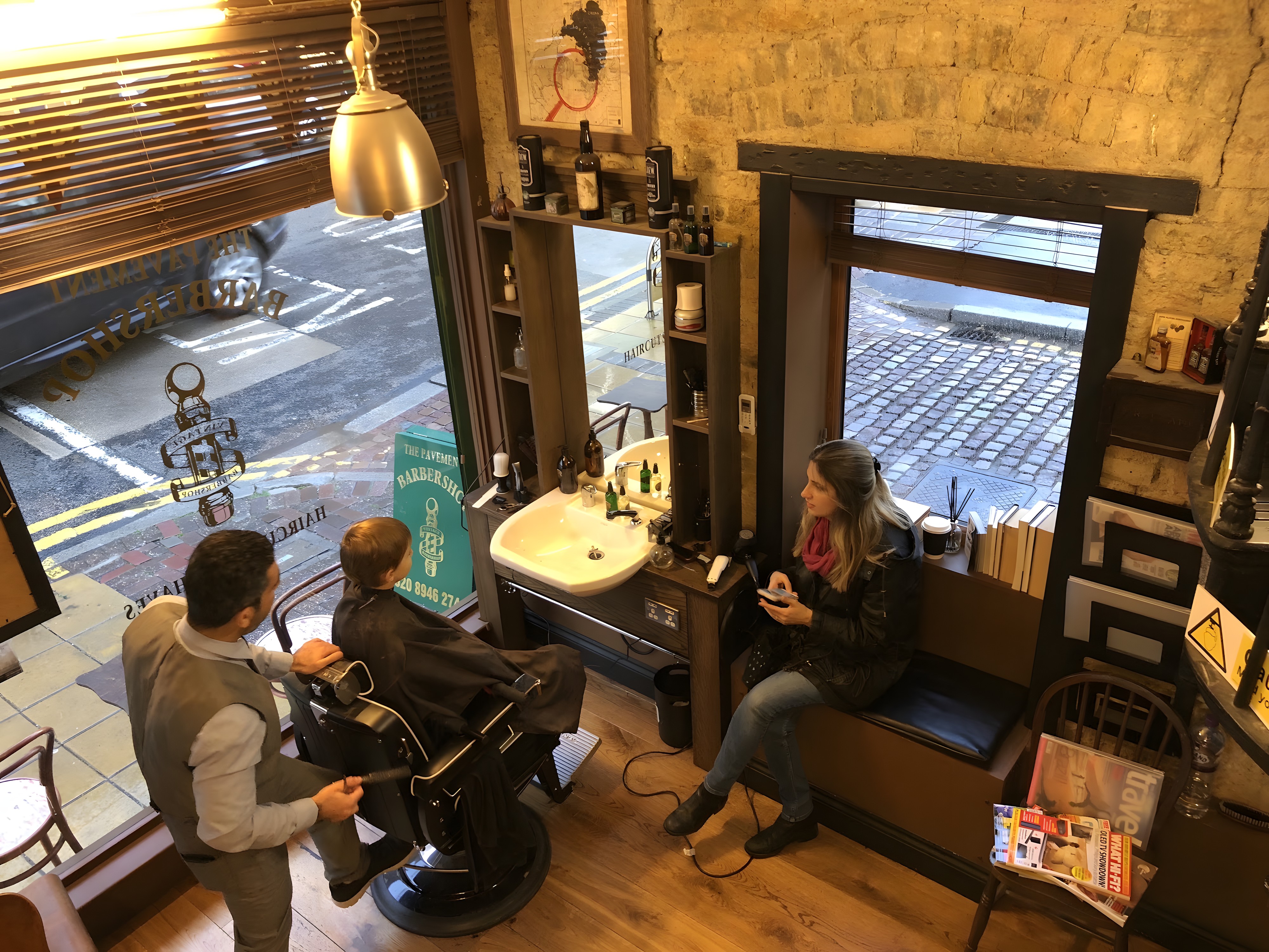 A barber at work by the window of The Pavement Barbershop, the shop name lettered in gold on the glass and the cobbled street beyond